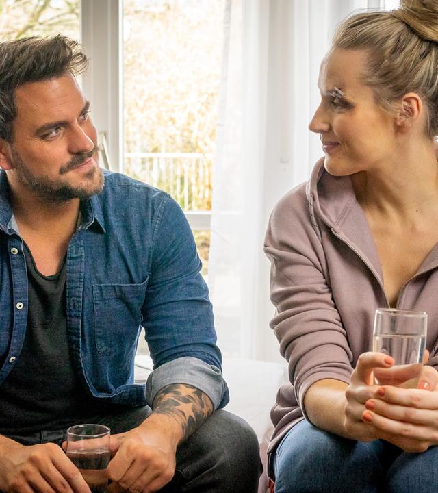 "Notruf Hafenkante - Neues Spiel, neues Glück": Kris (Marc Barthel) und Lena (Anna Hofbauer) sitzen nebeneinander auf einer Bettkante und lächeln sich an. Beide halten ein Glas Wasser in den Händen.