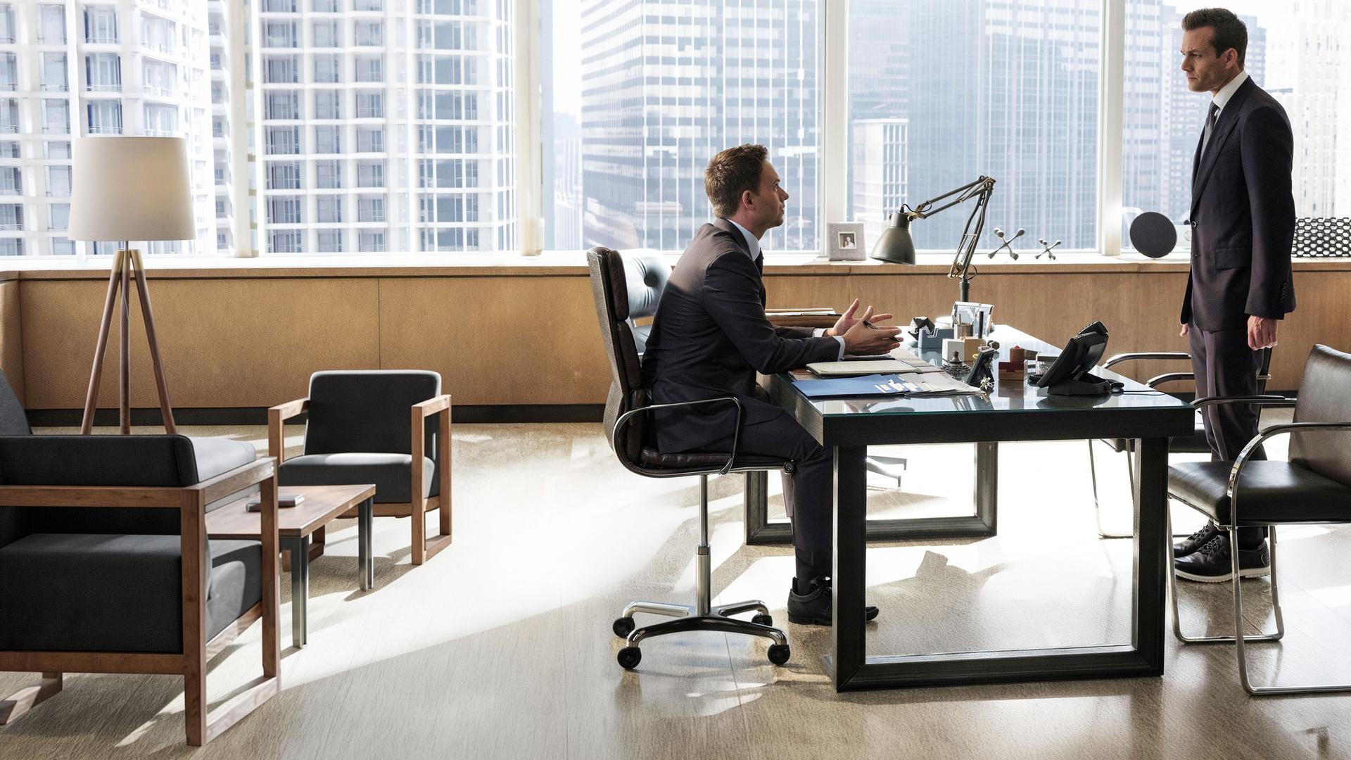 "Suits - Neuordnung": Mike (Patrick J. Adams) sitzt am Schreibtisch, beide Hände auf dem Tisch abgelegt. Er gestikuliert dabei und schaut den vor ihm stehenden Harvey (Gabriel Macht) eindringlich an. Durch die Fensterfront sind im Hintergrund Hochhäuser New Yorks zu erkennen.