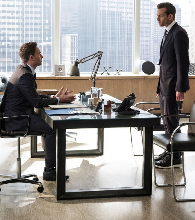 "Suits - Neuordnung": Mike (Patrick J. Adams) sitzt am Schreibtisch, beide Hände auf dem Tisch abgelegt. Er gestikuliert dabei und schaut den vor ihm stehenden Harvey (Gabriel Macht) eindringlich an. Durch die Fensterfront sind im Hintergrund Hochhäuser New Yorks zu erkennen.