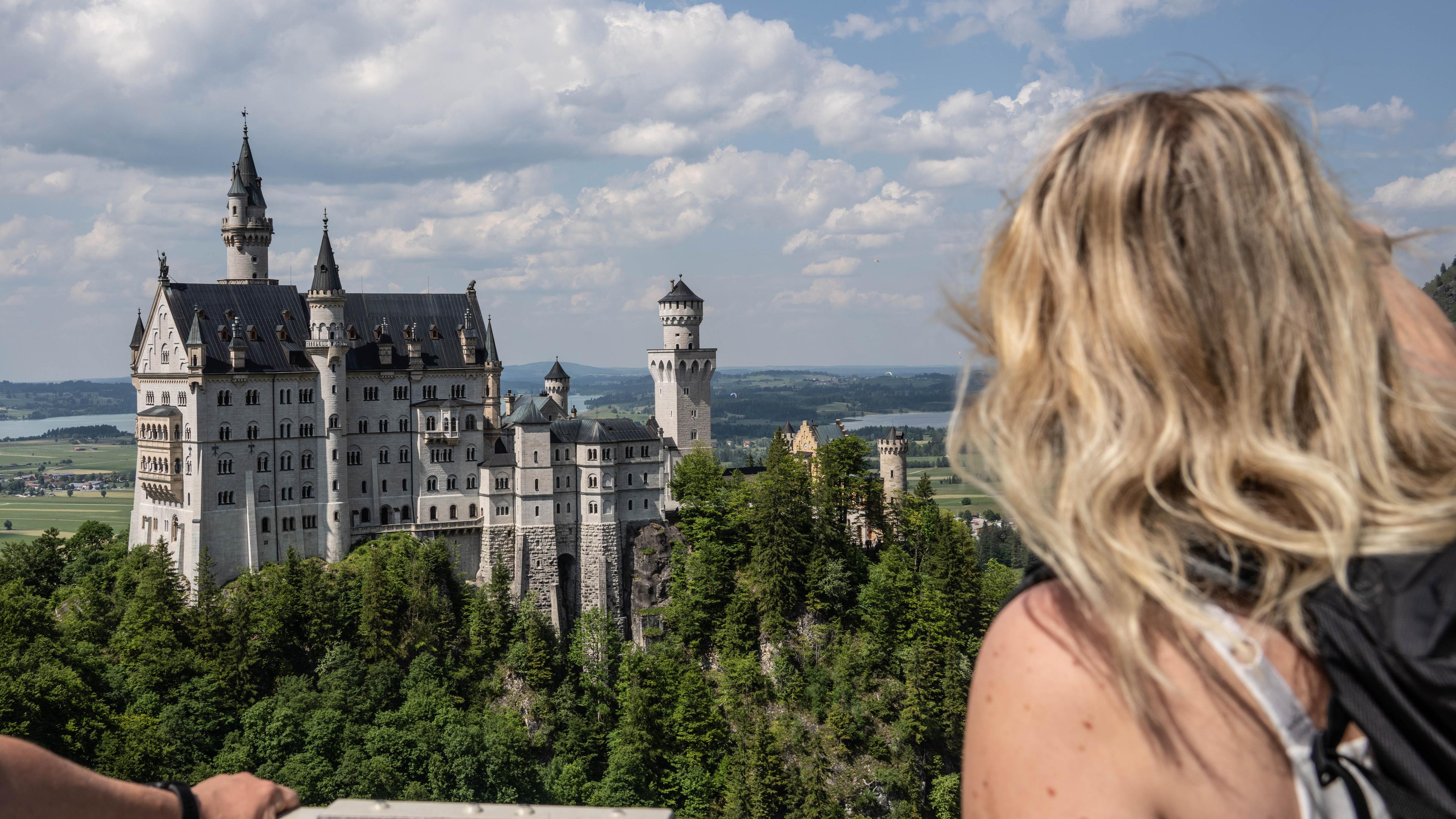 Zu sehen ist eine Frau, die von einer Hängebrücke aus auf Schloss Neuschwanstein blickt. 