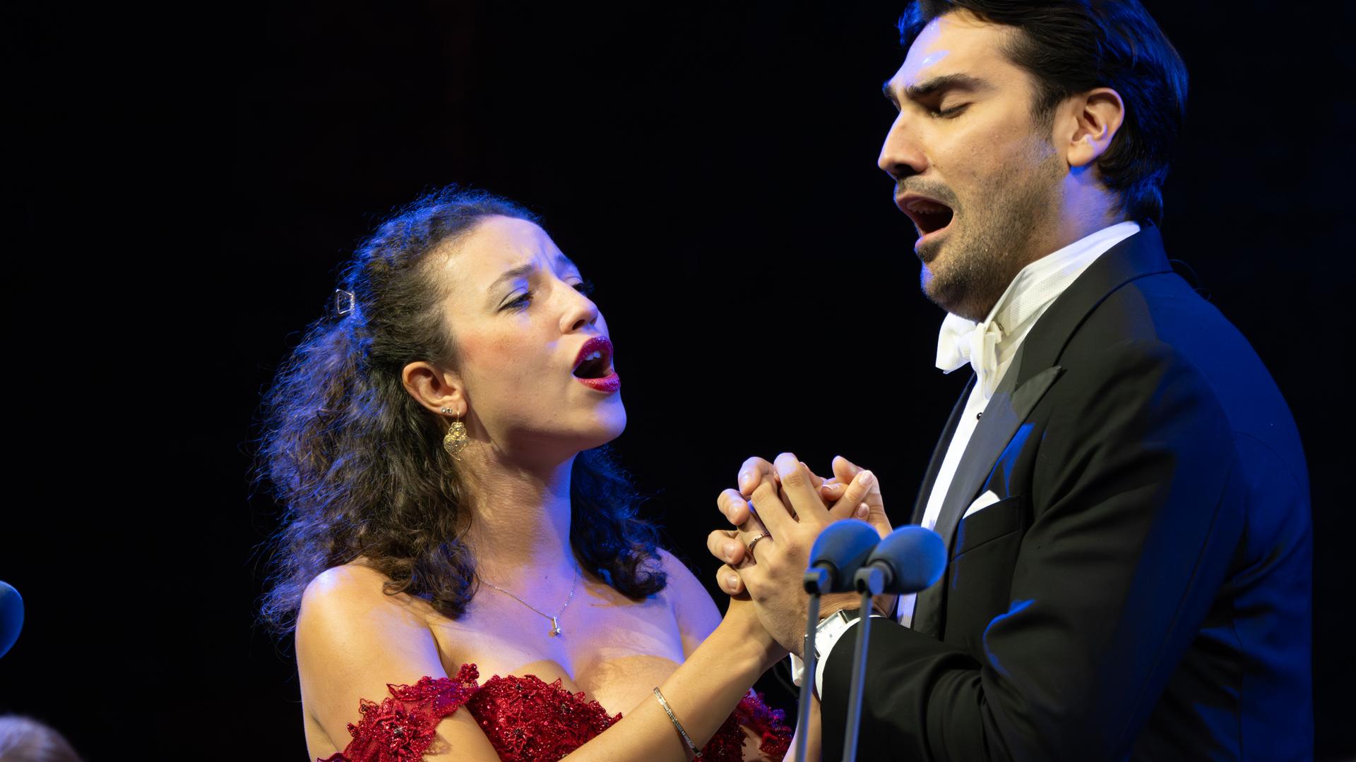 Jonathan Tetelman und Chelsea Zurflüh singen ein Duett. Dabei sind sie sich zugewandt und halten sich an den Händen. Zurflüh trägt ein rotes, schulterfreies Kleid und Tetelman trägt einen Smoking.