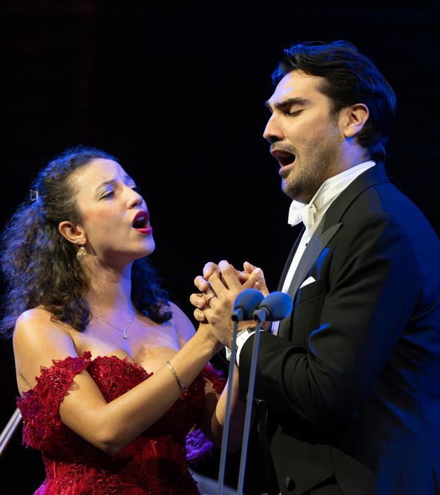 Jonathan Tetelman und Chelsea Zurflüh singen ein Duett. Dabei sind sie sich zugewandt und halten sich an den Händen. Zurflüh trägt ein rotes, schulterfreies Kleid und Tetelman trägt einen Smoking.