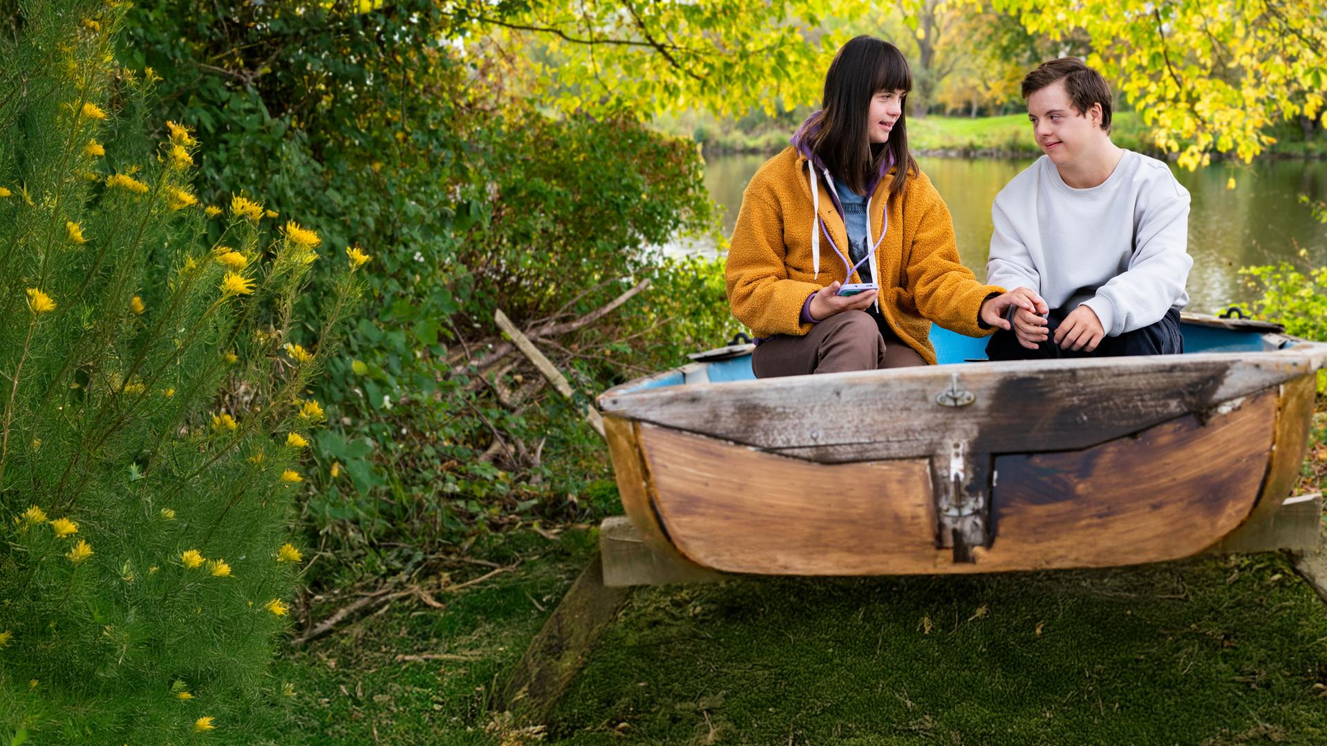 Emma (Juliane Siebecke) und Felix (Benjamin Raue) sitzen in einem alten Boot, das am Ufer eines Gewässers liegt, umgeben von vielen Bäumen und Sträuchern.