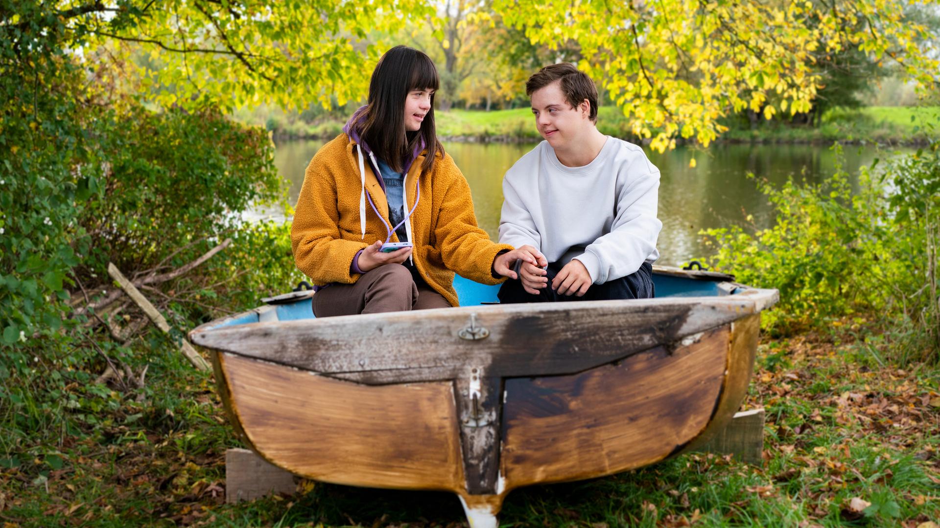 Emma (Juliane Siebecke) und Felix (Benjamin Raue) sitzen in einem alten Boot, das am Ufer eines Gewässers liegt, umgeben von vielen Bäumen und Sträuchern.