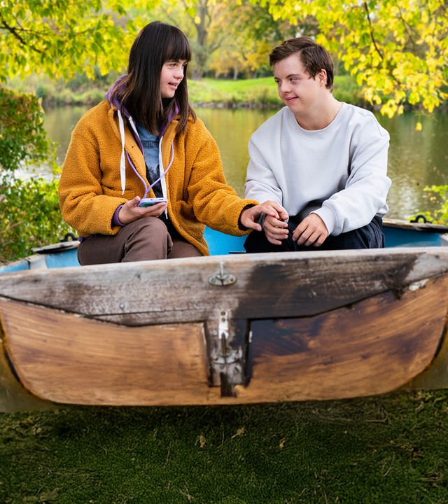 Emma (Juliane Siebecke) und Felix (Benjamin Raue) sitzen in einem alten Boot, das am Ufer eines Gewässers liegt, umgeben von vielen Bäumen und Sträuchern.