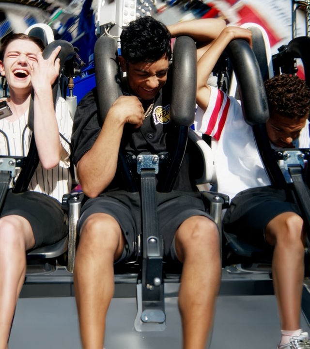 "After Benjamin: Neverland": Espen (Magnus Johan Tenfjord), Omer (Benjamin Ahmed) und Jama G (Nickolai Sødal Kabanda) sitzen nebeneinander in einer fahrenden Achterbahn. Sie halten sich an den Sicherheitsbügeln fest und haben sichtlich Spaß.