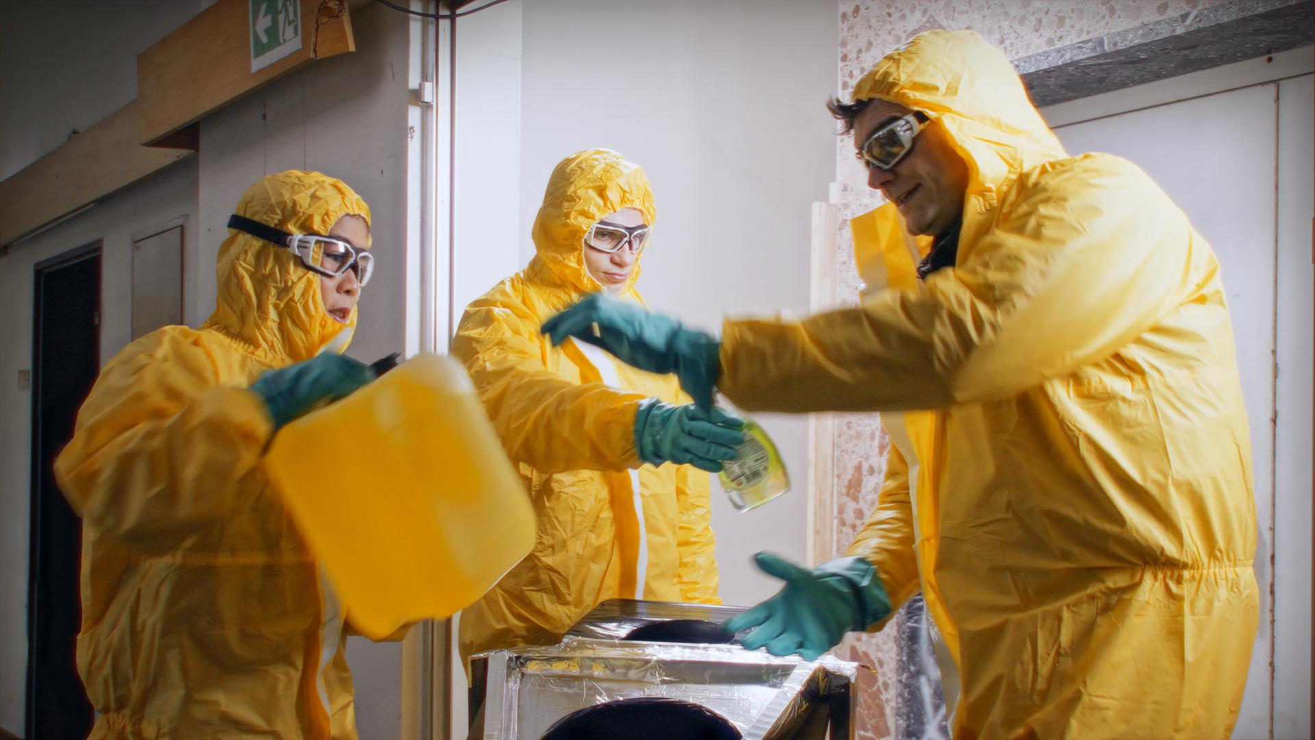Mai Thi Nguyen-Kim, Lutz van der Horst und Fabian Köster tragen Schutzbrillen und gelbe Schutzanzüge und hantieren mit gelben Kanistern. 
