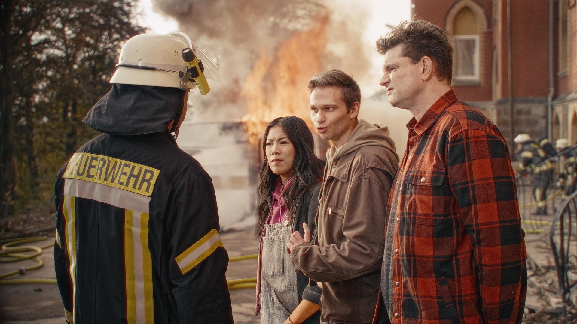 Ein Feuerwehrmann steht links im Bild mit dem Rücken zu Kamera und schaut die drei Moderatoren Mai Thi Nguyen-Kim, Fabian Köster und Lutz van der Horst (v.l.n.) an.  