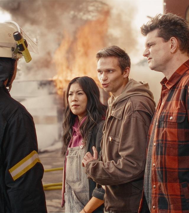 Ein Feuerwehrmann steht links im Bild mit dem Rücken zu Kamera und schaut die drei Moderatoren Mai Thi Nguyen-Kim, Fabian Köster und Lutz van der Horst (v.l.n.) an.  