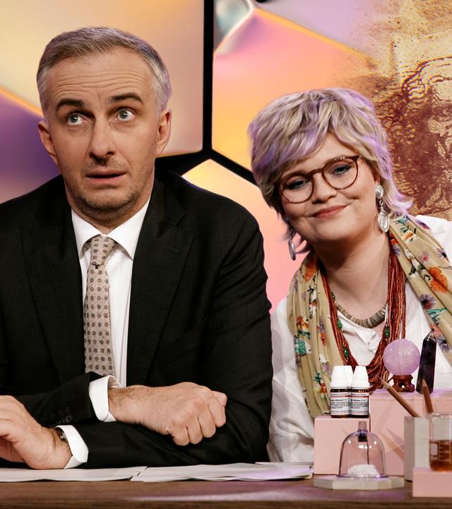 Jan Böhmermann sitzt am Schreibtisch. Neben ihm sitzt Carolin Worbs, die eine Kurzhaar-Perücke und eine Brille trägt und sich zu ihm hinüberlehnt.