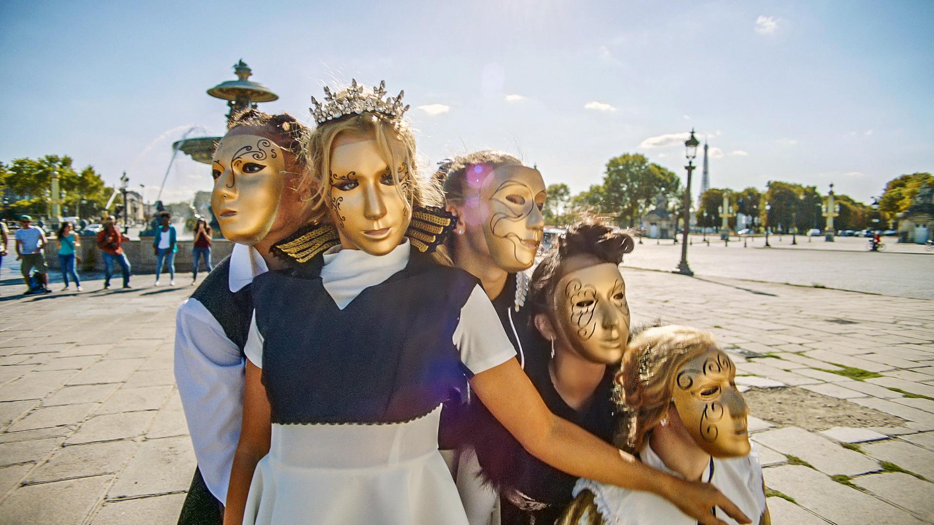 Fünf BLOK-Tänzerinnen stehen vor einem Brunnen auf einem Platz. Alle tragen goldene Masken und tragen Prinzessinnen-Kostüme.
