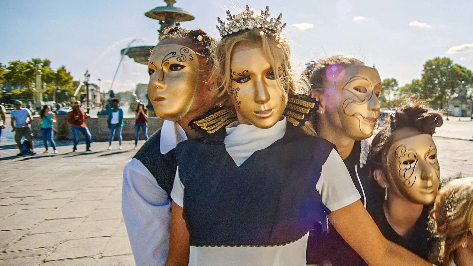 Fünf BLOK-Tänzerinnen stehen vor einem Brunnen auf einem Platz. Alle tragen goldene Masken und tragen Prinzessinnen-Kostüme.
