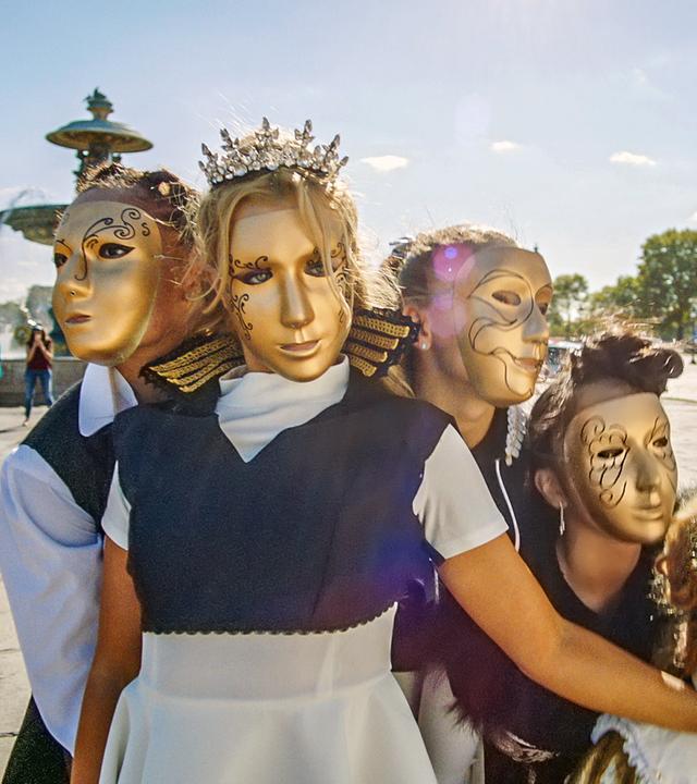 Fünf BLOK-Tänzerinnen stehen vor einem Brunnen auf einem Platz. Alle tragen goldene Masken und tragen Prinzessinnen-Kostüme.