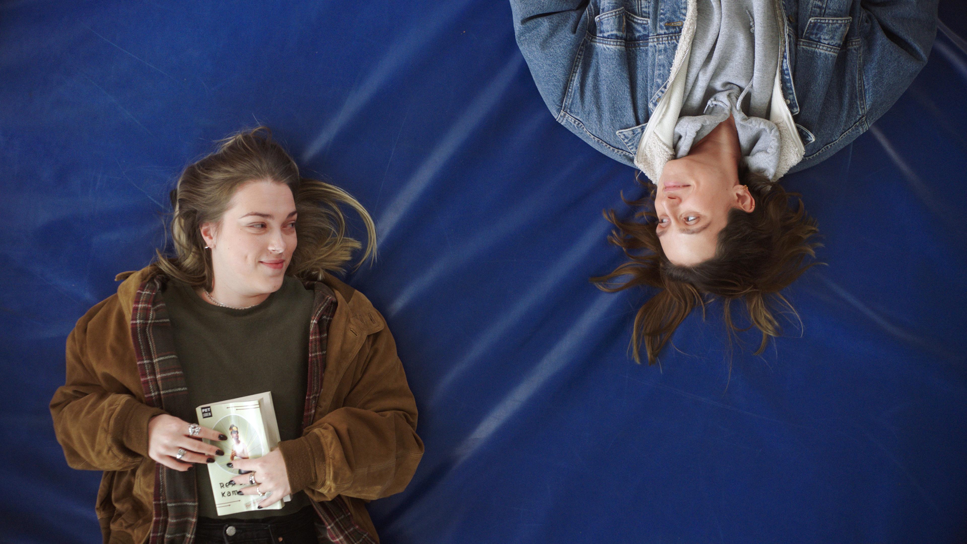 Man schaut von oben auf zwei junge Frauen, die auf einer blauen Turnmatte liegen und sich anschauen. Eine von ihnen hält ein Buch in den Händen.