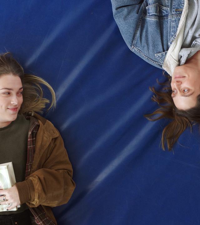 Man schaut von oben auf zwei junge Frauen, die auf einer blauen Turnmatte liegen und sich anschauen. Eine von ihnen hält ein Buch in den Händen.