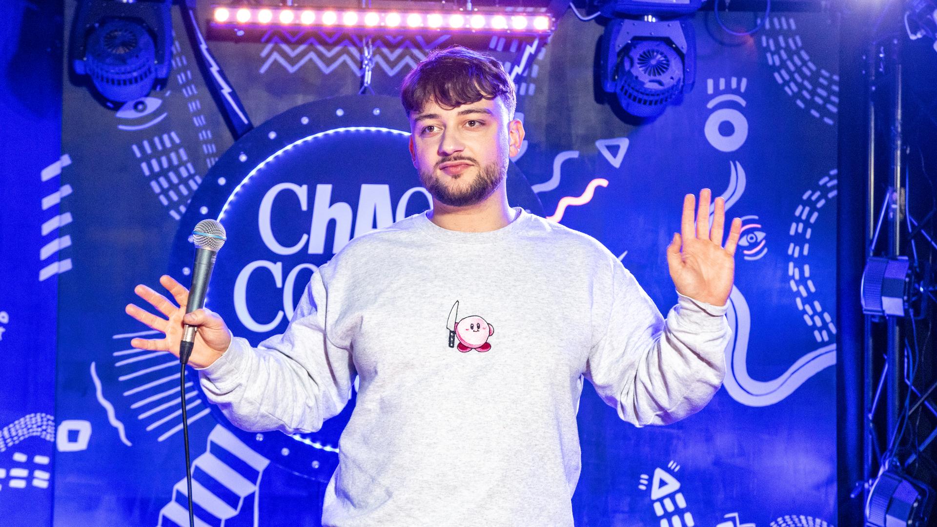 Niclas Amling steht mittig auf der Bühne. Er hält beide Hände seitlich weg von seinem Körper. Er trägt einen grauen Pullover.