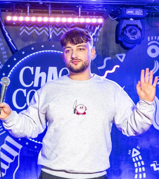 Niclas Amling steht mittig auf der Bühne. Er hält beide Hände seitlich weg von seinem Körper. Er trägt einen grauen Pullover.