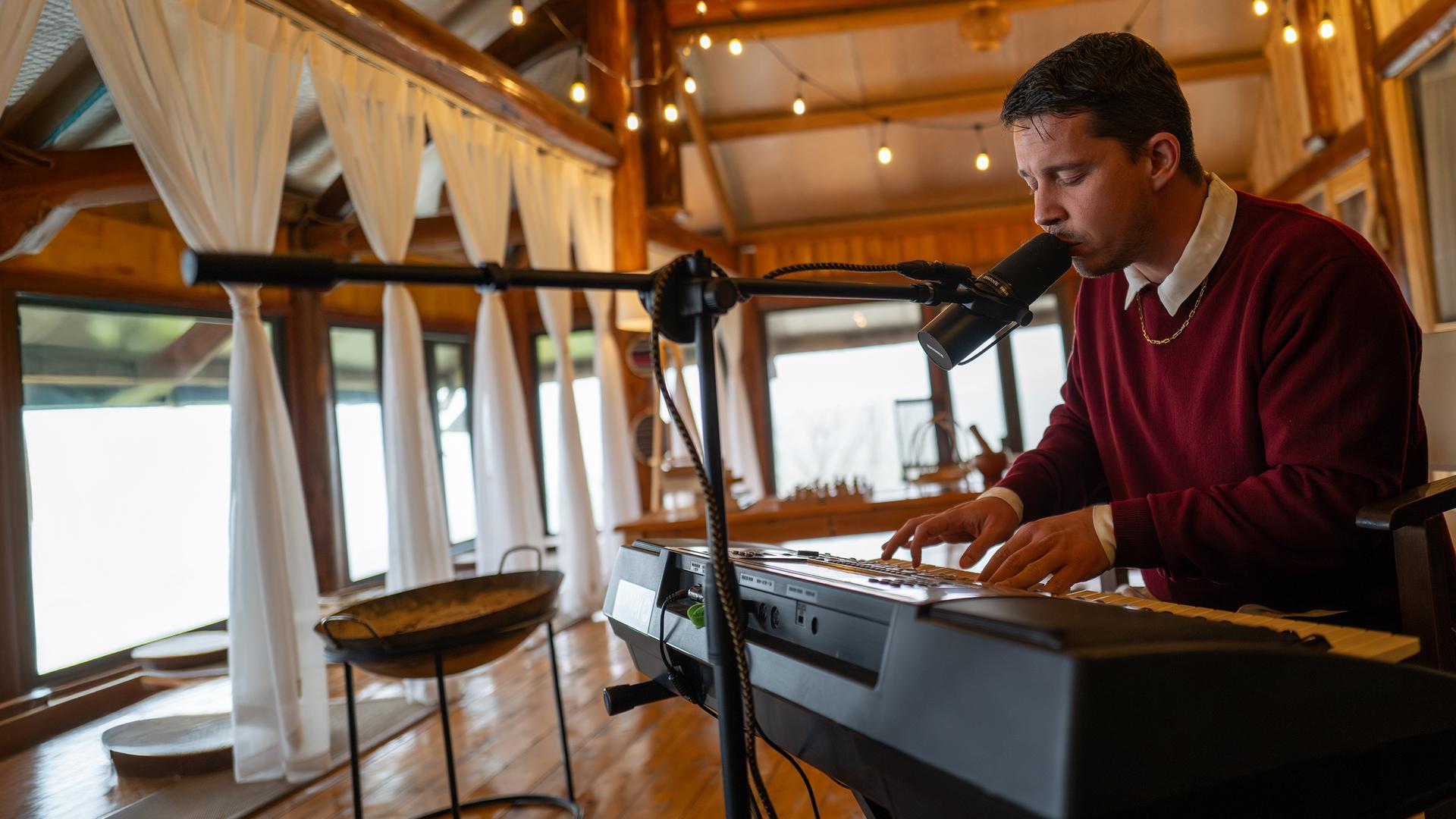 Nico Santos sitzt und spielt auf einem Keyboard und singt dabei in ein Mikrofon, das vor ihm steht. 