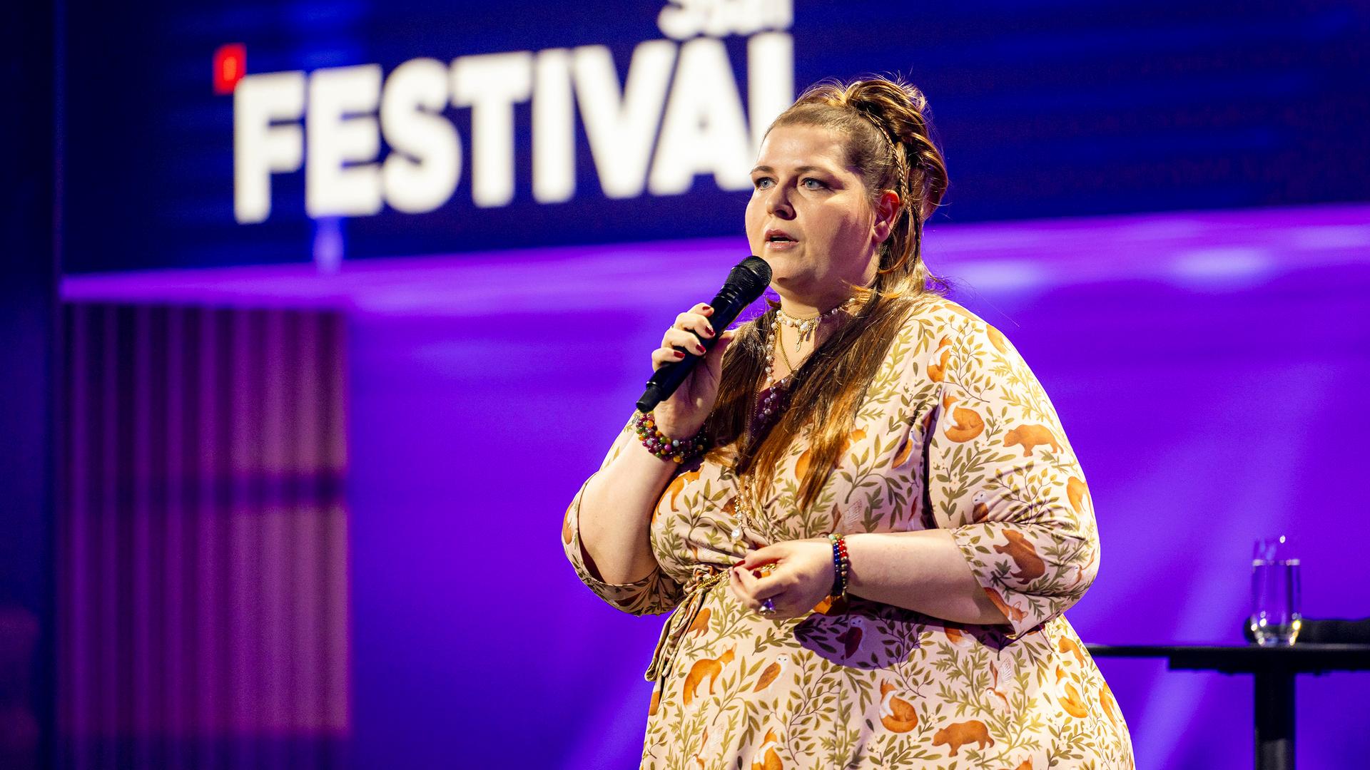Kabarettistin teht in der Mitte des Bildes. Sie blickt zum linken Bildrand. In ihrer rechten Hand hält sie ein Mikrofon. Sie trägt ein gemustertes Kleid. Die Bühne ist blau und lila beleuchtet.