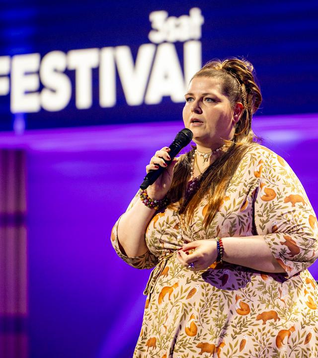 Kabarettistin teht in der Mitte des Bildes. Sie blickt zum linken Bildrand. In ihrer rechten Hand hält sie ein Mikrofon. Sie trägt ein gemustertes Kleid. Die Bühne ist blau und lila beleuchtet.