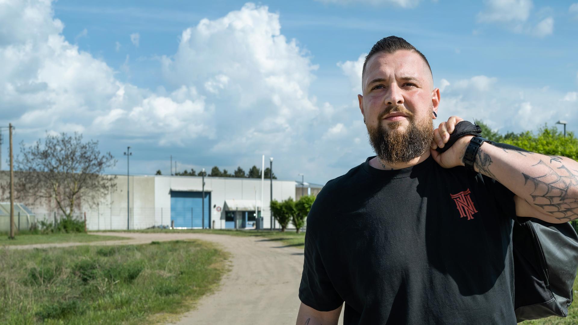 Alex (32) läuft auf die Kamera zu. Er trägt ein schwarzes T-Shirt und eine schwarze Hose. Sein linker Arm ist angewinkelt, er trägt eine Reisetasche um seine linke Schulter. Er geht einen Weg entlang, der von niedrigem Gras gesäumt ist. Im Hintergrund steht ein niedriges Gefängnisgebäude aus grauem Beton mit blauem Tor.