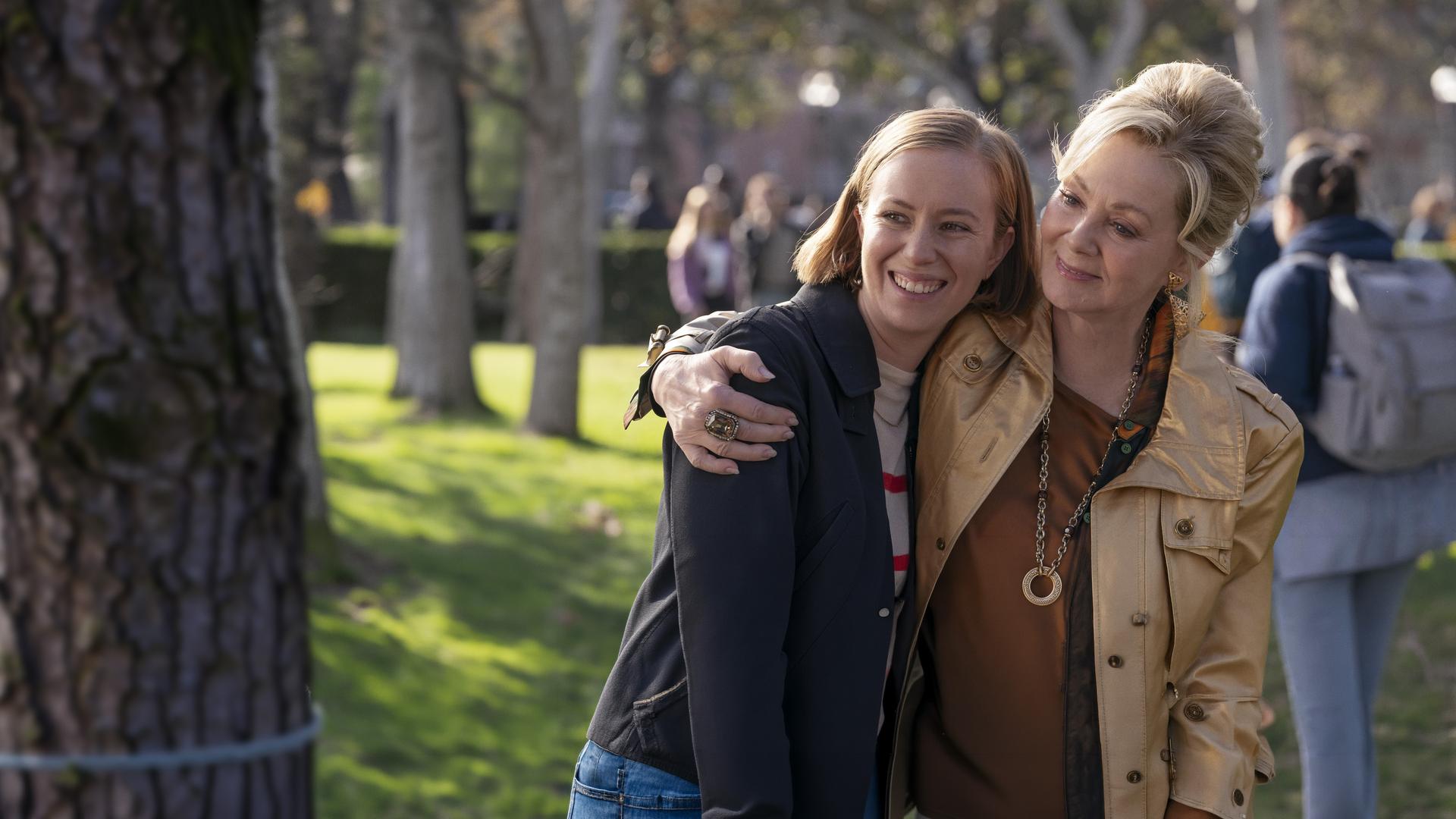 "Hacks - Niemals für einen Witz entschuldigen": Deborah (Jean Smart) lächelt und umarmt Ava Hannah Einbinder), die ebenso gelöst und glücklich lacht. Im Hintergrund befinden sich, unscharf zu erkennen, weitere Personen (Komparsen) unter Bäumen.
