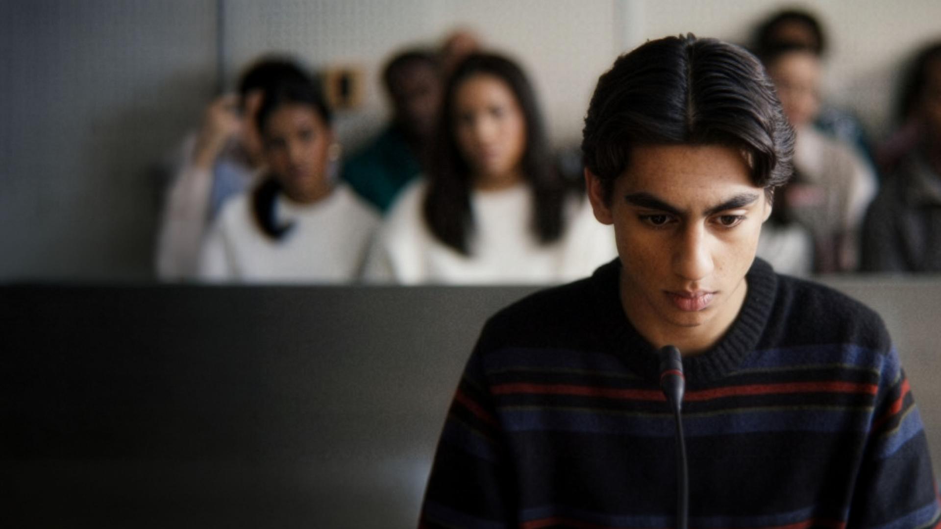 "After Benjamin: Niemals vergessen": Elias (Sam Ashraf) sitzt im Zeugenstand vor Gericht. Er blickt nachdenklich nach vorn, vor ihm steht ein Mikrofon. Im Hintergrund sitzen, unscharf zu erkennen, Zuschauer und Zuschauerinnen im Gerichtssaal.