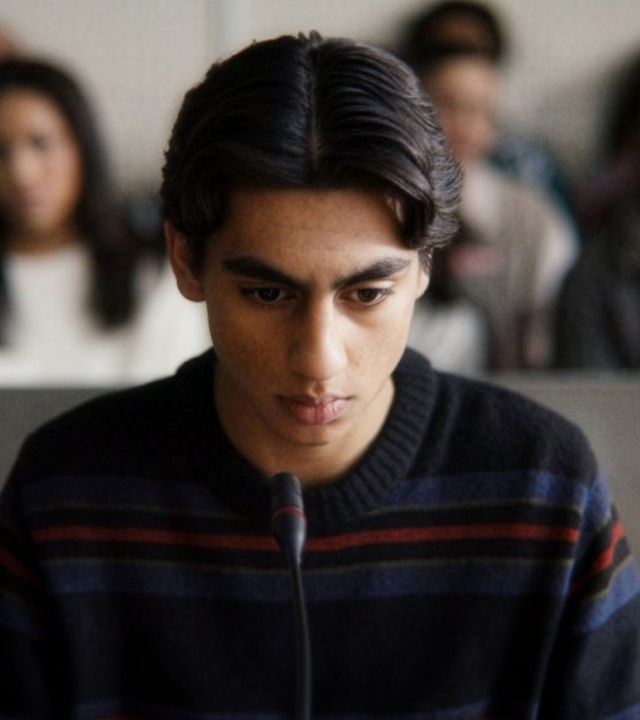 "After Benjamin: Niemals vergessen": Elias (Sam Ashraf) sitzt im Zeugenstand vor Gericht. Er blickt nachdenklich nach vorn, vor ihm steht ein Mikrofon. Im Hintergrund sitzen, unscharf zu erkennen, Zuschauer und Zuschauerinnen im Gerichtssaal.