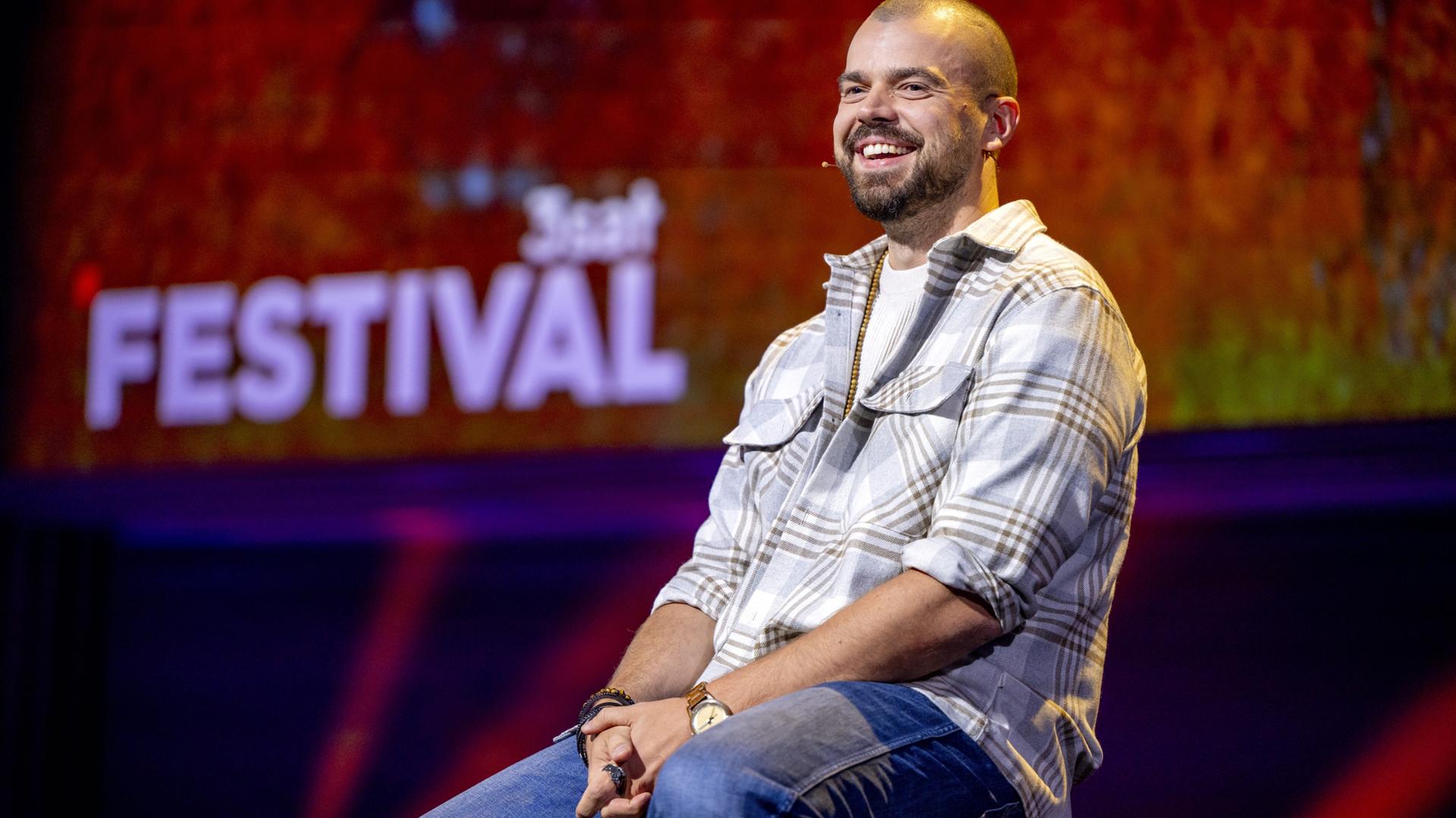 Nikita Miller sitzt auf einem Barhocker in der Mitte des Bildes. Er trägt ein offenes kariertes Hemd und lächelt. Der Hintergrund ist rot beleuchtet.