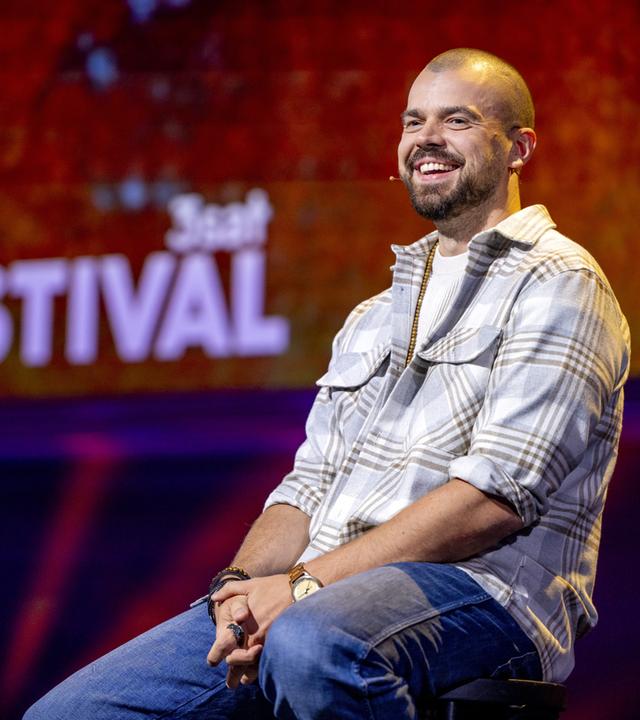 Nikita Miller sitzt auf einem Barhocker in der Mitte des Bildes. Er trägt ein offenes kariertes Hemd und lächelt. Der Hintergrund ist rot beleuchtet.