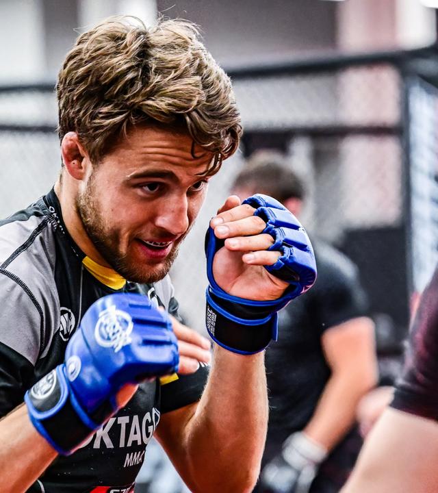 Ein junger Mann mit Bart, in Trainingskleidung mit Schutzhandschuhen, kämpft in einem Studio gegen einen Gegner. 