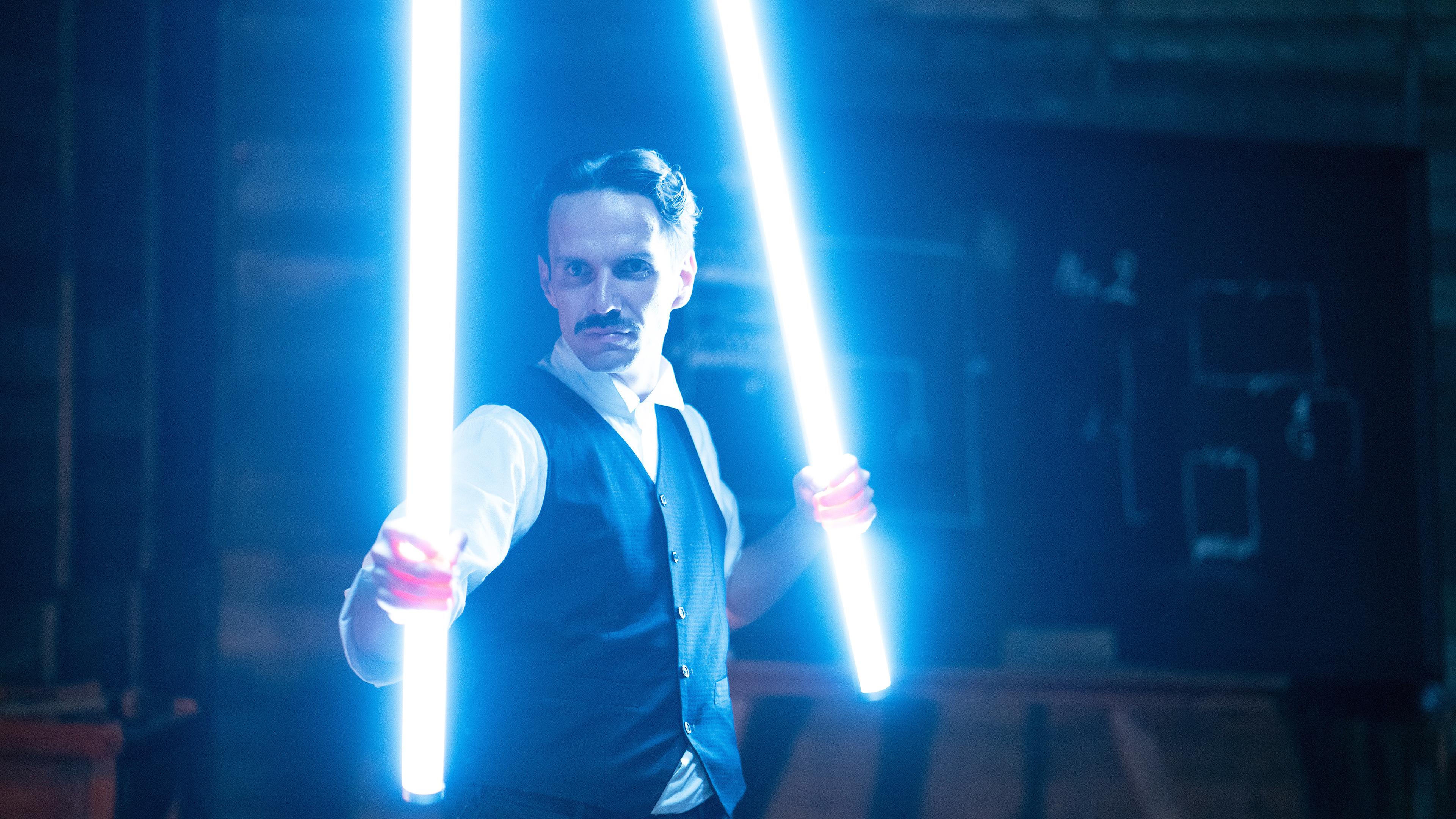 Spielszene: Nikola Tesla steht in seiner Werkstatt mit leuchtenden Röhren, die er wie Schwerter in der Hand hält.