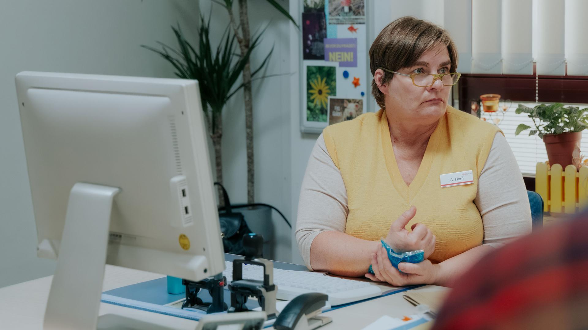 "Nine to Five - Das Amt - Ein Dachs": Gisela (Petra Hartung) sitzt an ihrem Schreibtisch und blickt zur Seite. Sie hÃ¤lt einen KÃ¼hlakku an ihre Hand. Hinter ihr sind die Fenster mit einer Jalousie geschlossenen, eine Pflanze steht davor auf dem Fensterbrett, neben dem Fenster hÃ¤ngt eine Magnettafel.