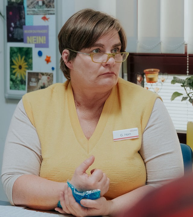 "Nine to Five - Das Amt - Ein Dachs": Gisela (Petra Hartung) sitzt an ihrem Schreibtisch und blickt zur Seite. Sie hÃ¤lt einen KÃ¼hlakku an ihre Hand. Hinter ihr sind die Fenster mit einer Jalousie geschlossenen, eine Pflanze steht davor auf dem Fensterbrett, neben dem Fenster hÃ¤ngt eine Magnettafel.