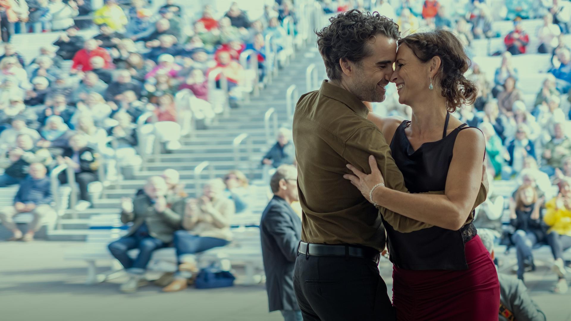 Ina Behrendsen (Julia Brendler) schmiegt sich an ihren Kollegen Hinnerk Feldmann (Oliver Wnuk). Die beiden tanzen lachend und eng umschlungen auf einer Bühne im Freien. Im Hintergrund sitzt ein buntes Publikum auf den Rängen, vor der Bühne steht ein Musiker mit einem Kontrabass.