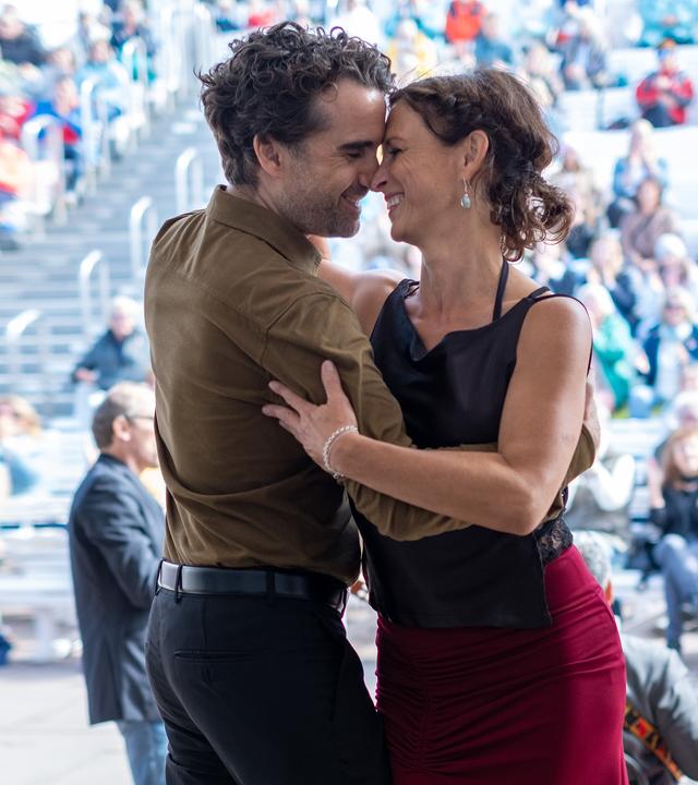 Ina Behrendsen (Julia Brendler) schmiegt sich an ihren Kollegen Hinnerk Feldmann (Oliver Wnuk). Die beiden tanzen lachend und eng umschlungen auf einer Bühne im Freien. Im Hintergrund sitzt ein buntes Publikum auf den Rängen, vor der Bühne steht ein Musiker mit einem Kontrabass.