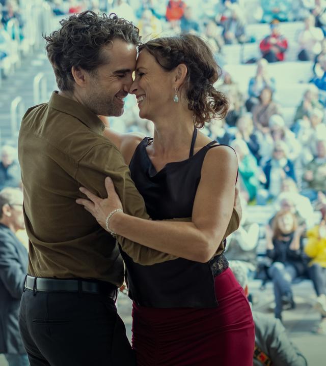 Ina Behrendsen (Julia Brendler) schmiegt sich an ihren Kollegen Hinnerk Feldmann (Oliver Wnuk). Die beiden tanzen lachend und eng umschlungen auf einer Bühne im Freien. Im Hintergrund sitzt ein buntes Publikum auf den Rängen, vor der Bühne steht ein Musiker mit einem Kontrabass.
