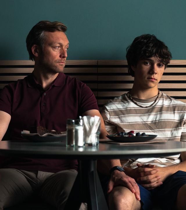 "No Dogs Allowed": Gabo (Carlo Krammling) und Dave (Robin Sondermann) sitzen nebeneinander an einem Cafétisch. Dave sieht zu Gabo und hat seine Hand auf dessen Oberschenkel gelegt. Gabo schaut gedankenverloren vor sich hin.