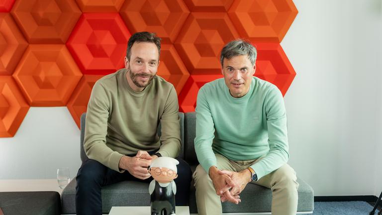 OMR-Podcast-Host Philipp Westermeyer (l.) im Gespräch mit ZDF-Intendant Norbert Himmler. Beide sitzen sich über Eck in jeweils einem Loungemöbel gegenüber. Im Hintergrund steht dazwischenm ein Mainzelmännchen