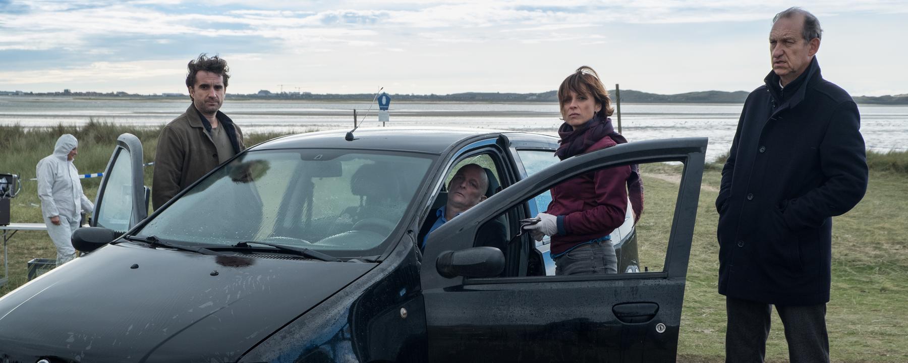 "Nord Nord Mord - Sievers und der schönste Tag": Hinter dem Steuer eines schwarzen Pkw sitzt der tote Hochzeitsvideograf Stefan Borg (Johnny West). Carl Sievers (Peter Heinrich Brix), Ina Behrendsen (Julia Brendler) und Hinnerk Feldmann (Oliver Wnuk ) stehen um das Auto herum. Im Hintergrund sind ein Kollege (Komparse) im weißen Overall und das Wattenmeer zu sehen. Der Himmel ist leicht bewölkt und es ist windig.