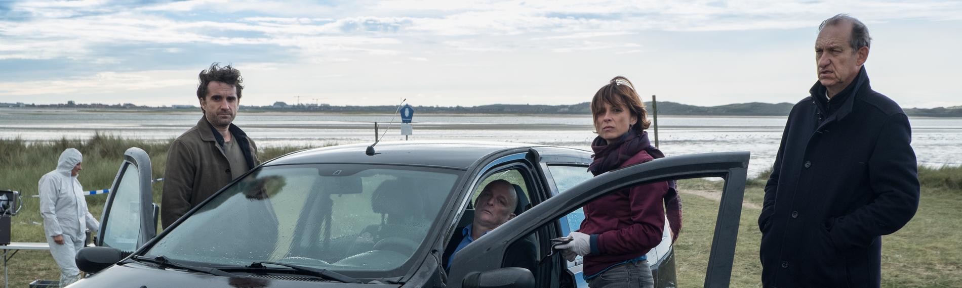 "Nord Nord Mord - Sievers und der schönste Tag": Hinter dem Steuer eines schwarzen Pkw sitzt der tote Hochzeitsvideograf Stefan Borg (Johnny West). Carl Sievers (Peter Heinrich Brix), Ina Behrendsen (Julia Brendler) und Hinnerk Feldmann (Oliver Wnuk ) stehen um das Auto herum. Im Hintergrund sind ein Kollege (Komparse) im weißen Overall und das Wattenmeer zu sehen. Der Himmel ist leicht bewölkt und es ist windig.