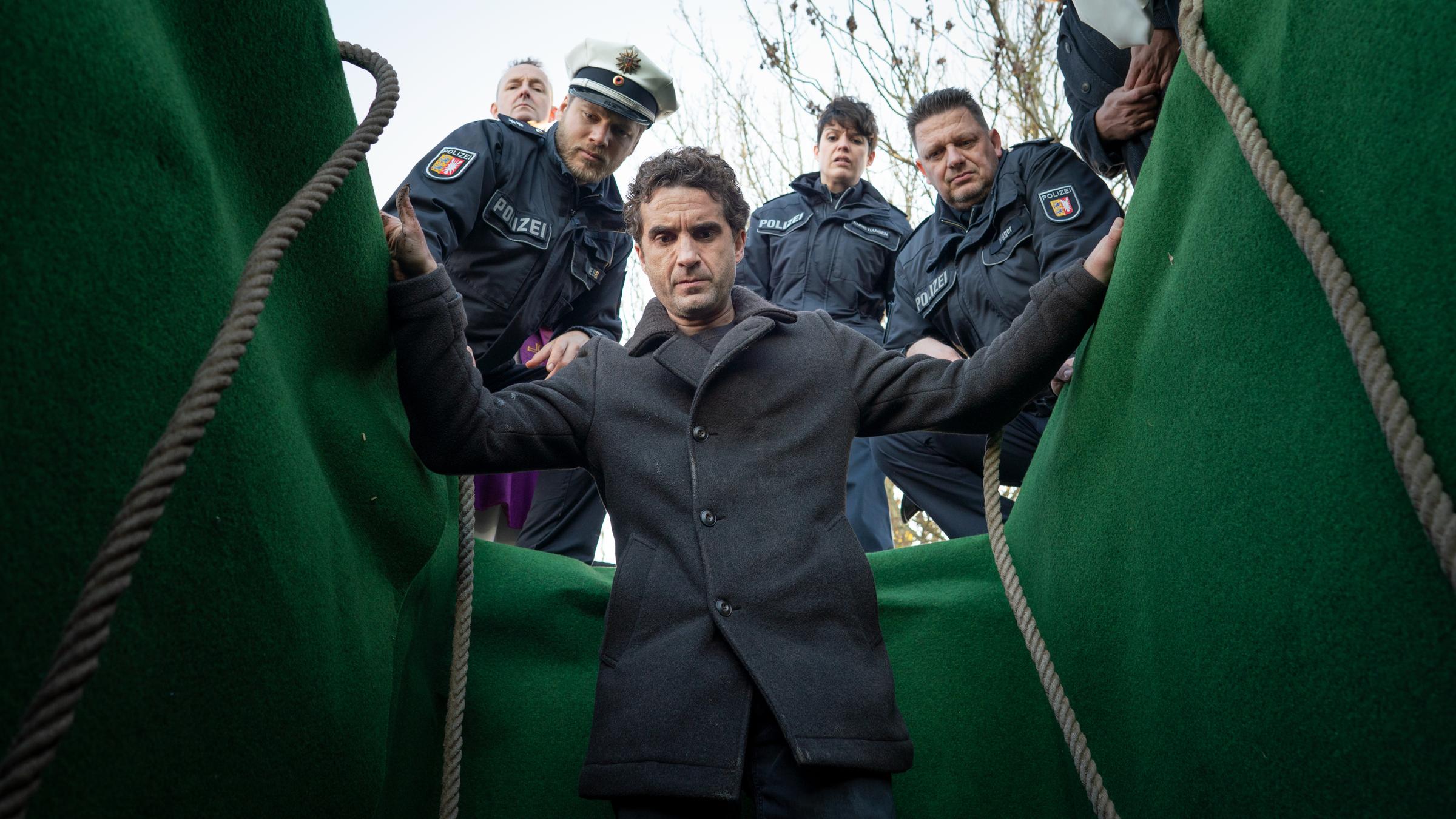 "Nord Nord Mord - Sievers und die letzte Beichte": Hinnerk Feldmann steht in einem offenen, mit grünem Tuch ausgeschlagenen Grab. Er hält sich mit den Händen am oberen Rand fest und schaut vor sich nach unten in die Tiefe. Hinter ihm am oberen Rand der Grube stehen uniformierte Polizisten und schauen ebenfalls nach vorne in das Grab hinein.