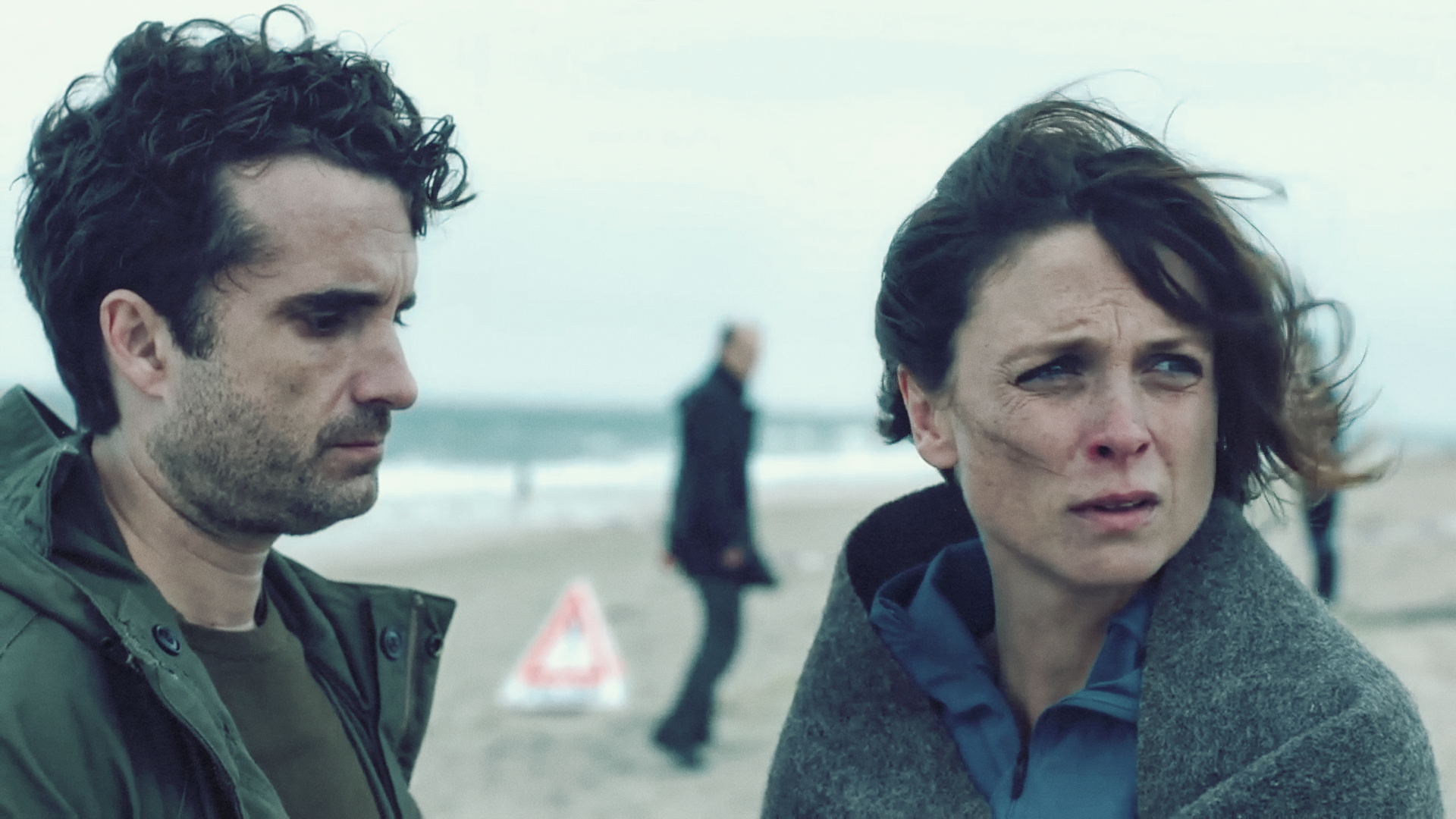 Hinnerk Feldmann schaut betreten zu Boden. Ihm gegenüber steht Ina Behrendsen, die sich in eine Schutzdecke gehüllt unsicher umsieht. Im Hintergurnd der Strand mit einem Warndreieck und ein paar Menschen, die den Tatort sichern.