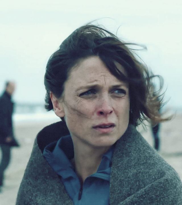 Hinnerk Feldmann schaut betreten zu Boden. Ihm gegenüber steht Ina Behrendsen, die sich in eine Schutzdecke gehüllt unsicher umsieht. Im Hintergurnd der Strand mit einem Warndreieck und ein paar Menschen, die den Tatort sichern.