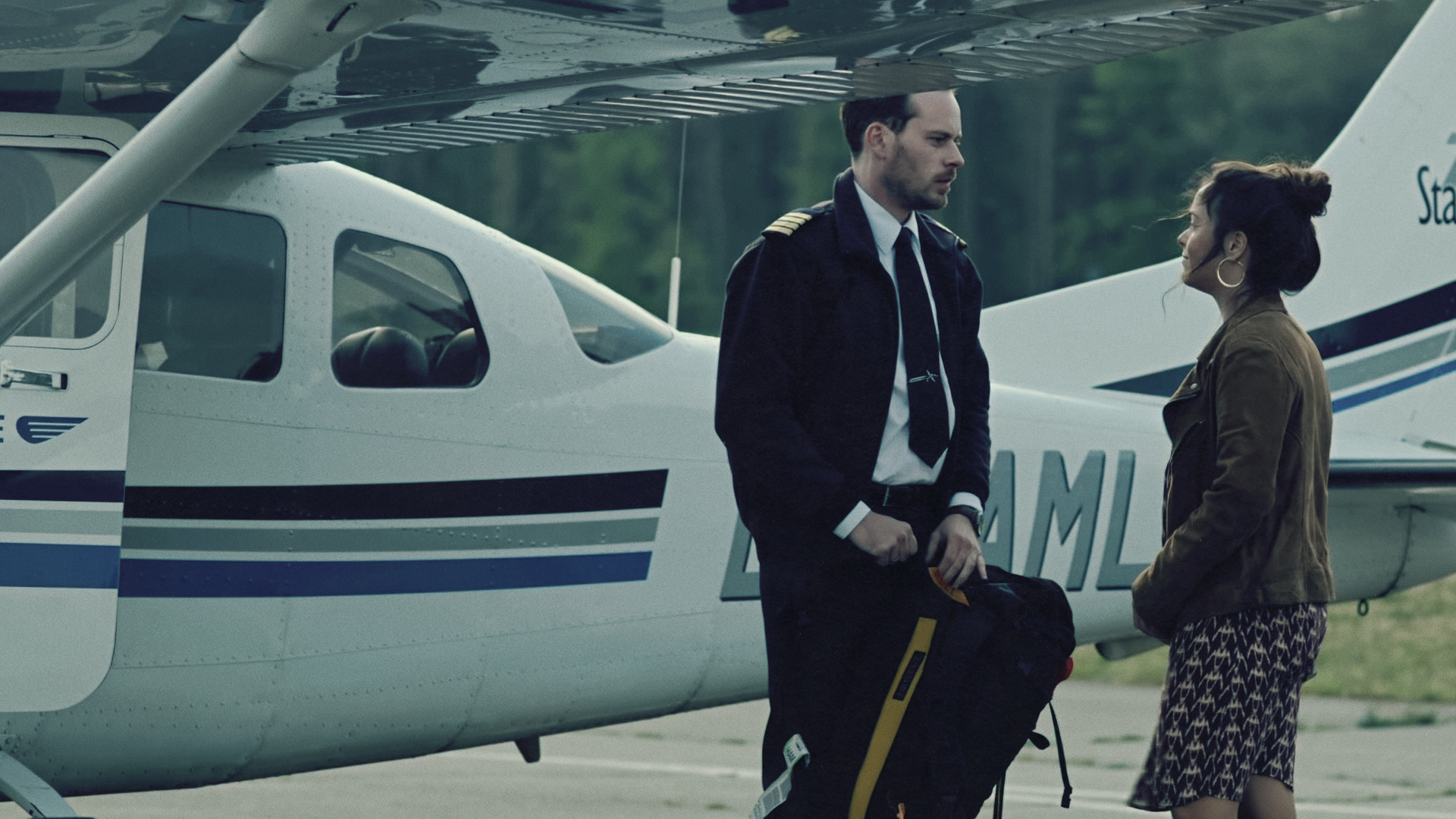 Ein Mann in Pilotenuniform und eine Frau stehen vor einem kleinen Privatflugzeug und schauen sich an.
