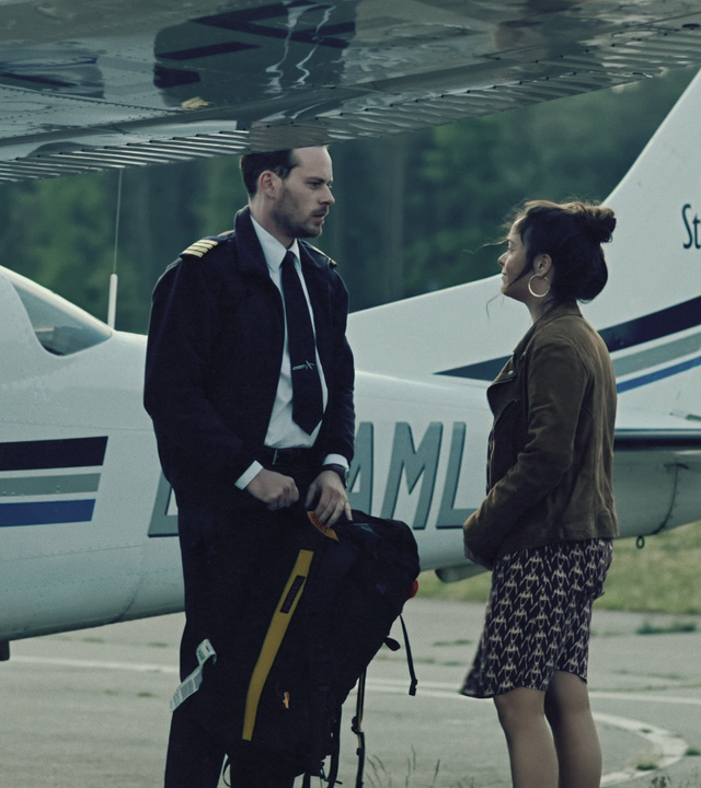 Ein Mann in Pilotenuniform und eine Frau stehen vor einem kleinen Privatflugzeug und schauen sich an.