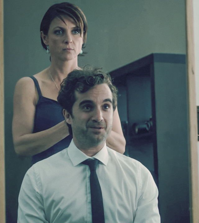 Ein Mann in Hemd und Kravatte sitzt vor einem Spiegel. Eine Frau im Kleid steht hinter ihm und schaut kritisch.