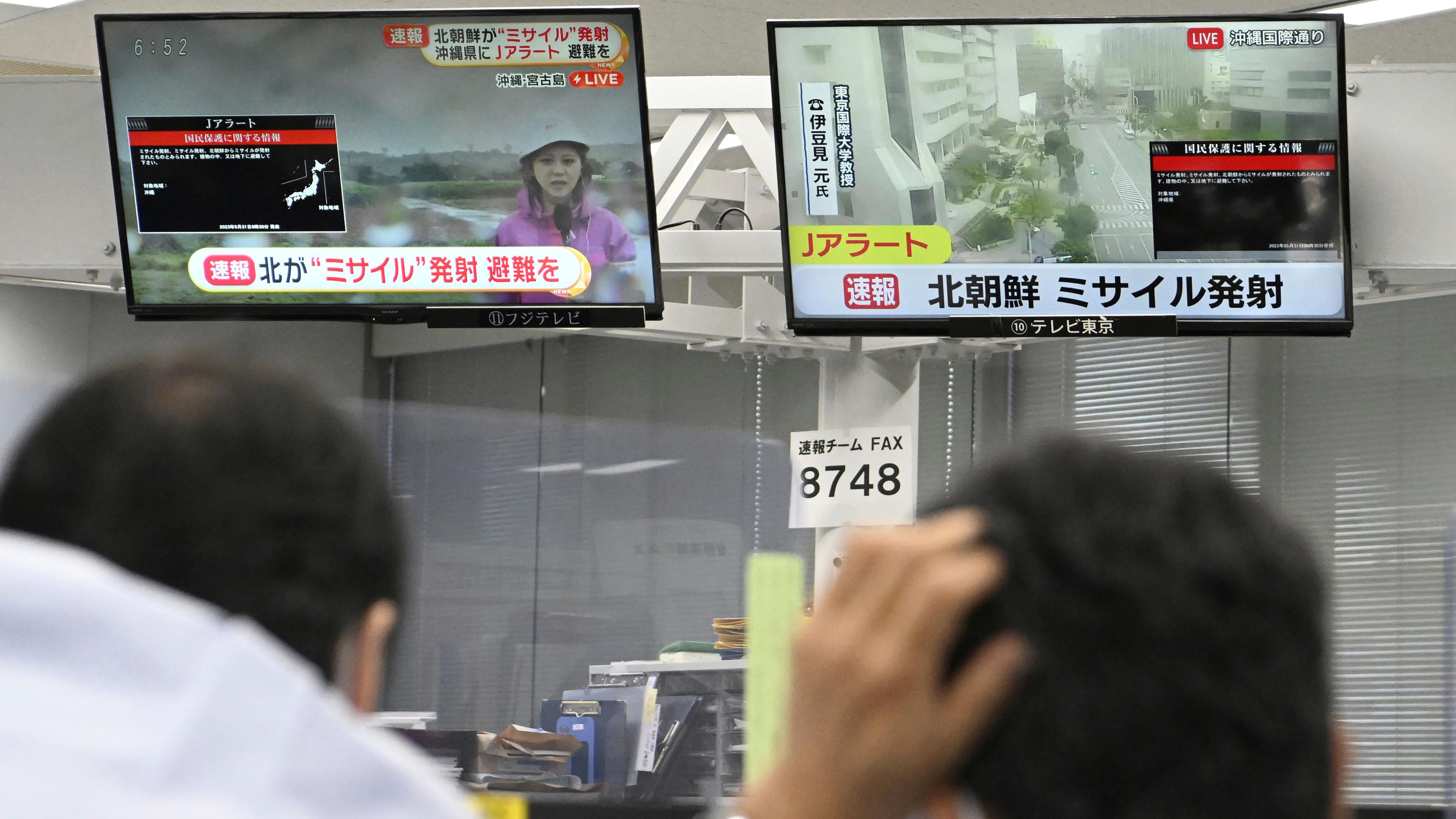 Zwei Männer schauen auf zwei Bildschirme die TV-Nachrichten zeigen. 