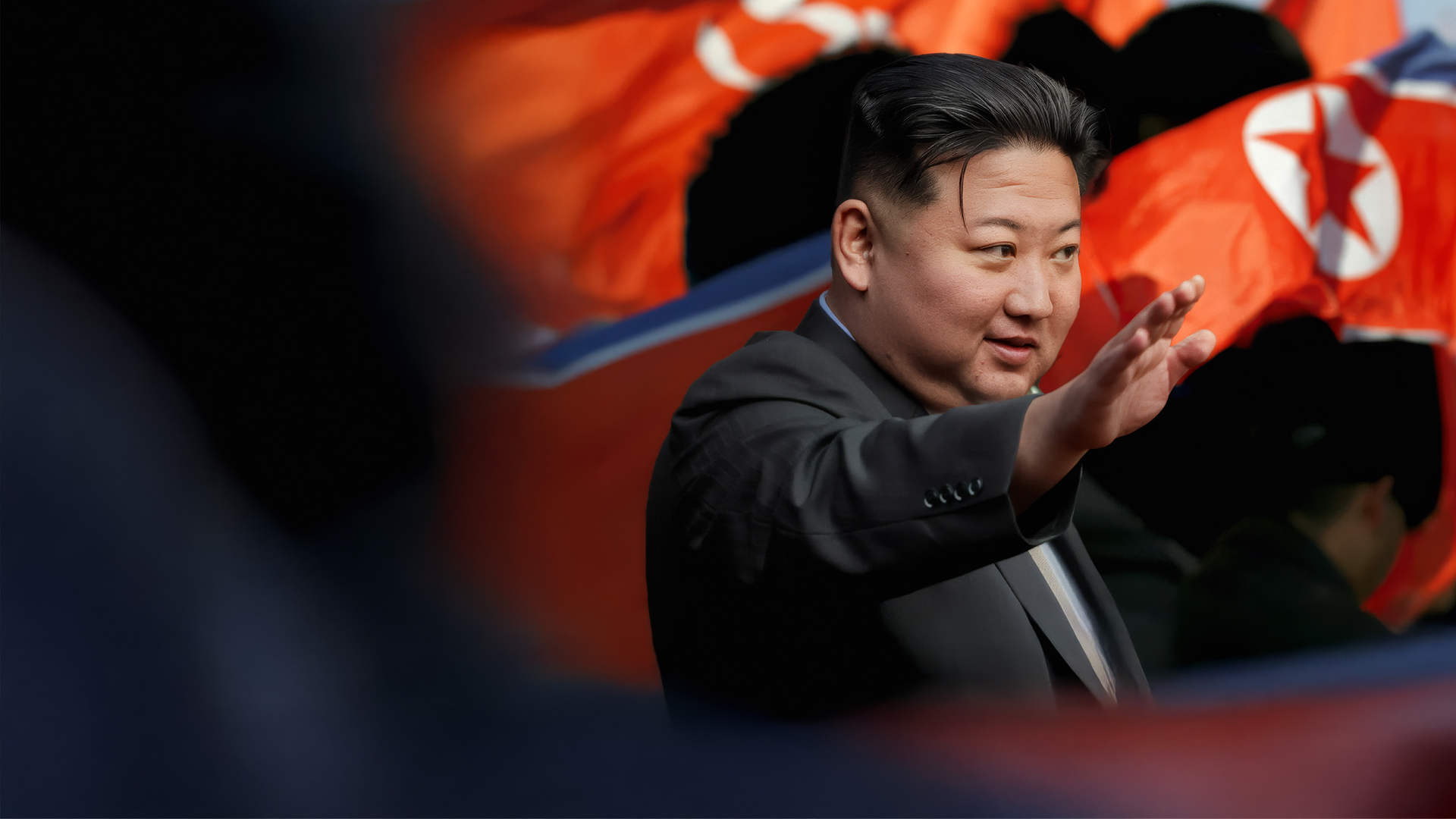 Kim Jong-un trägt einen schwarzen Anzug und streckt den rechten Arm nach vorne. Im Hintergrund wehen nordkoreanische Flaggen.
