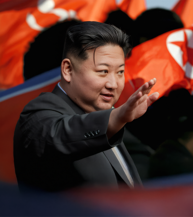 Kim Jong-un trägt einen schwarzen Anzug und streckt den rechten Arm nach vorne. Im Hintergrund wehen nordkoreanische Flaggen.
